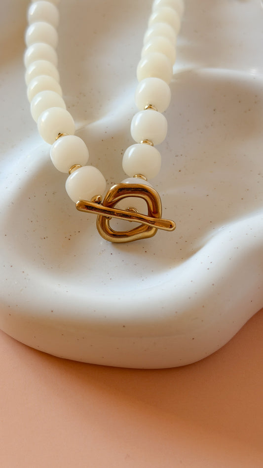 A close-up of a beaded necklace with round off-white beads and a gold toggle clasp, displayed on a sculptural white ceramic stand against a peach background.
