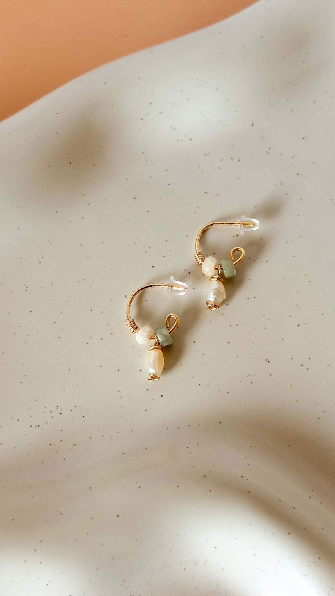 A pair of gold hoop earrings with white pearl drops and small light green bead accents, displayed on a textured off-white surface with soft shadows.