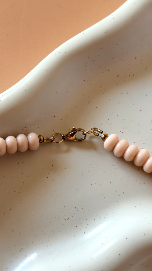A close-up of a beaded necklace with pale pink round beads and a gold clasp, resting on a white speckled ceramic dish against a warm beige background.