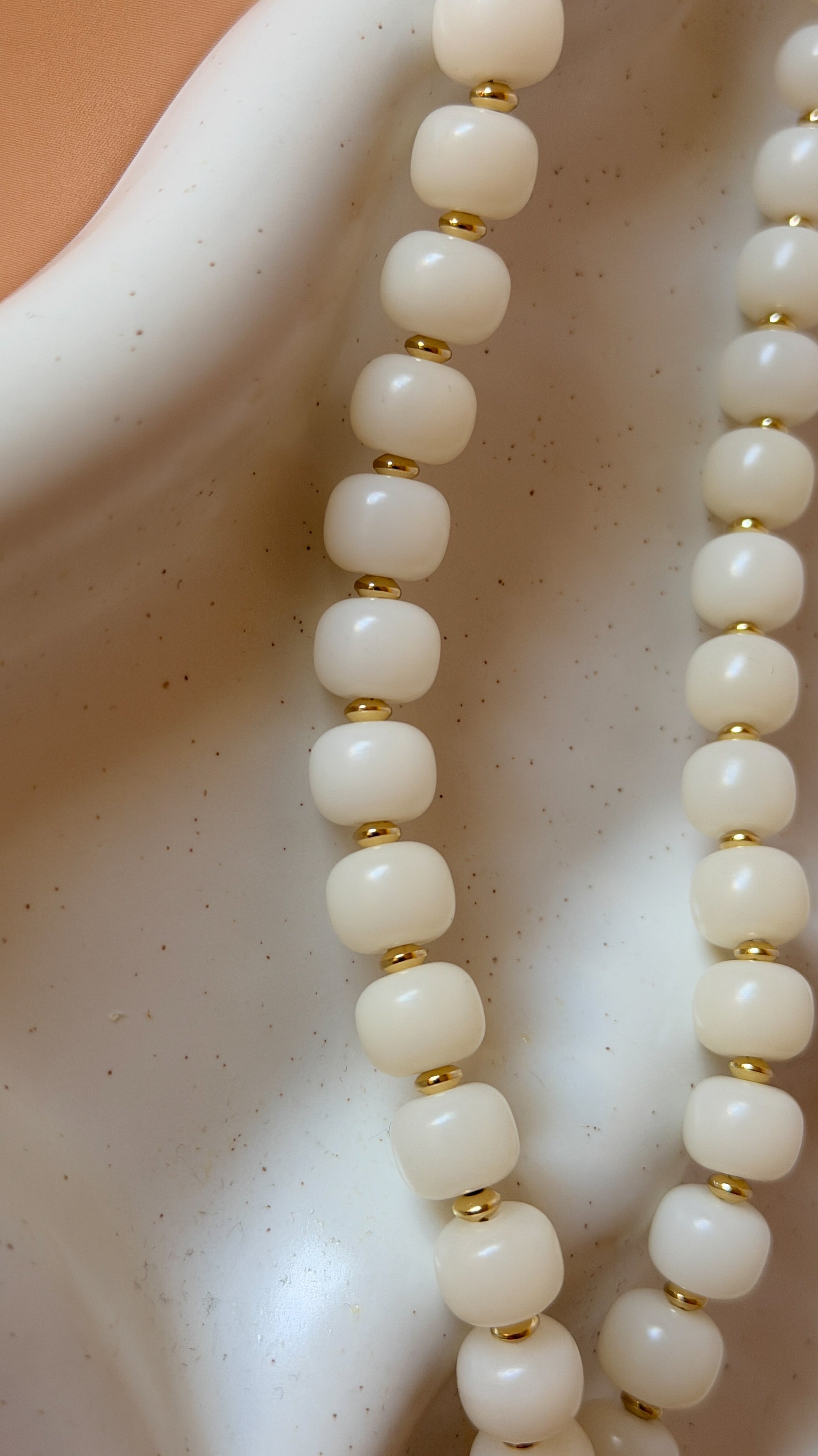A close-up of a beaded necklace with round off-white beads separated by small gold spacer beads, draped over a white ceramic display stand.