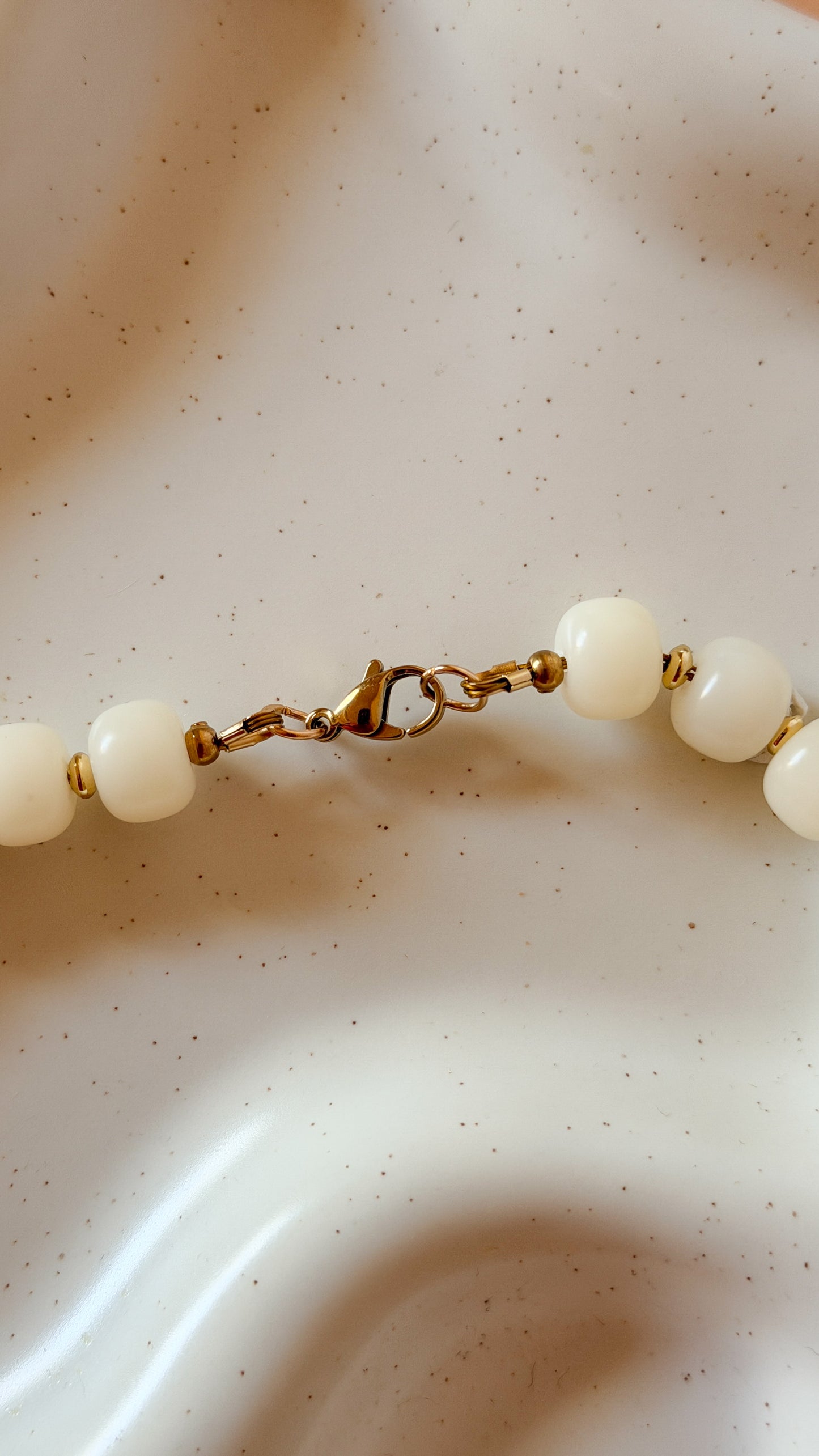 A close-up of a beaded necklace with round white beads and gold-toned metal links and clasp, resting on a textured off-white surface.