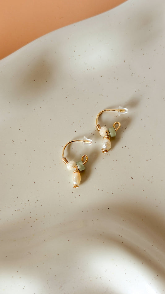A pair of gold hoop earrings with white pearl drops and small light green bead accents, displayed on a textured off-white surface with soft shadows.