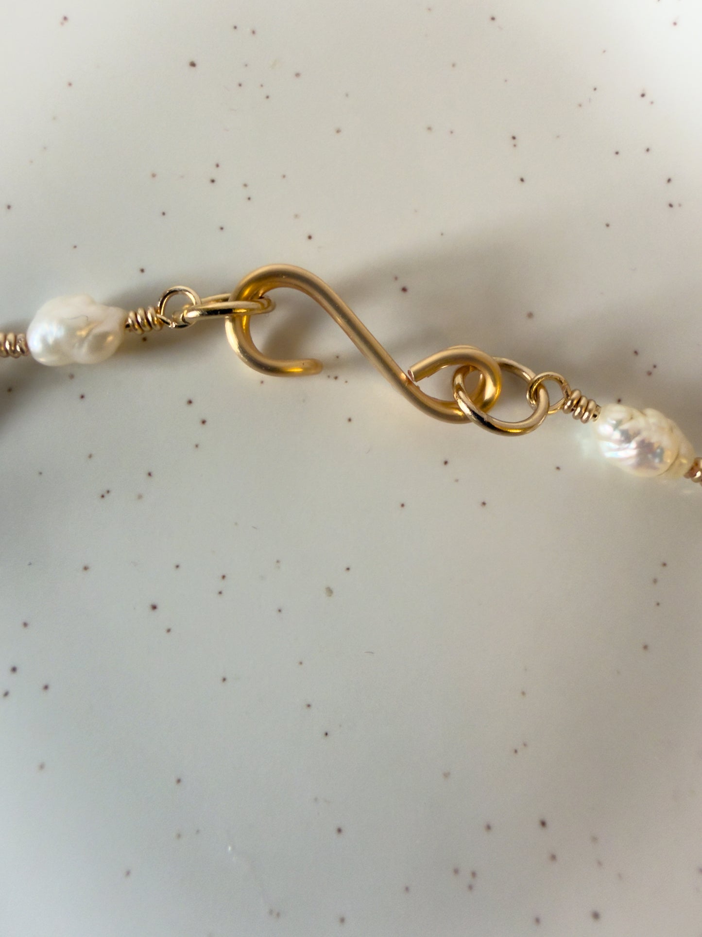 Close-up of a handmade gold necklace closure with pearl beads on a speckled beige background
