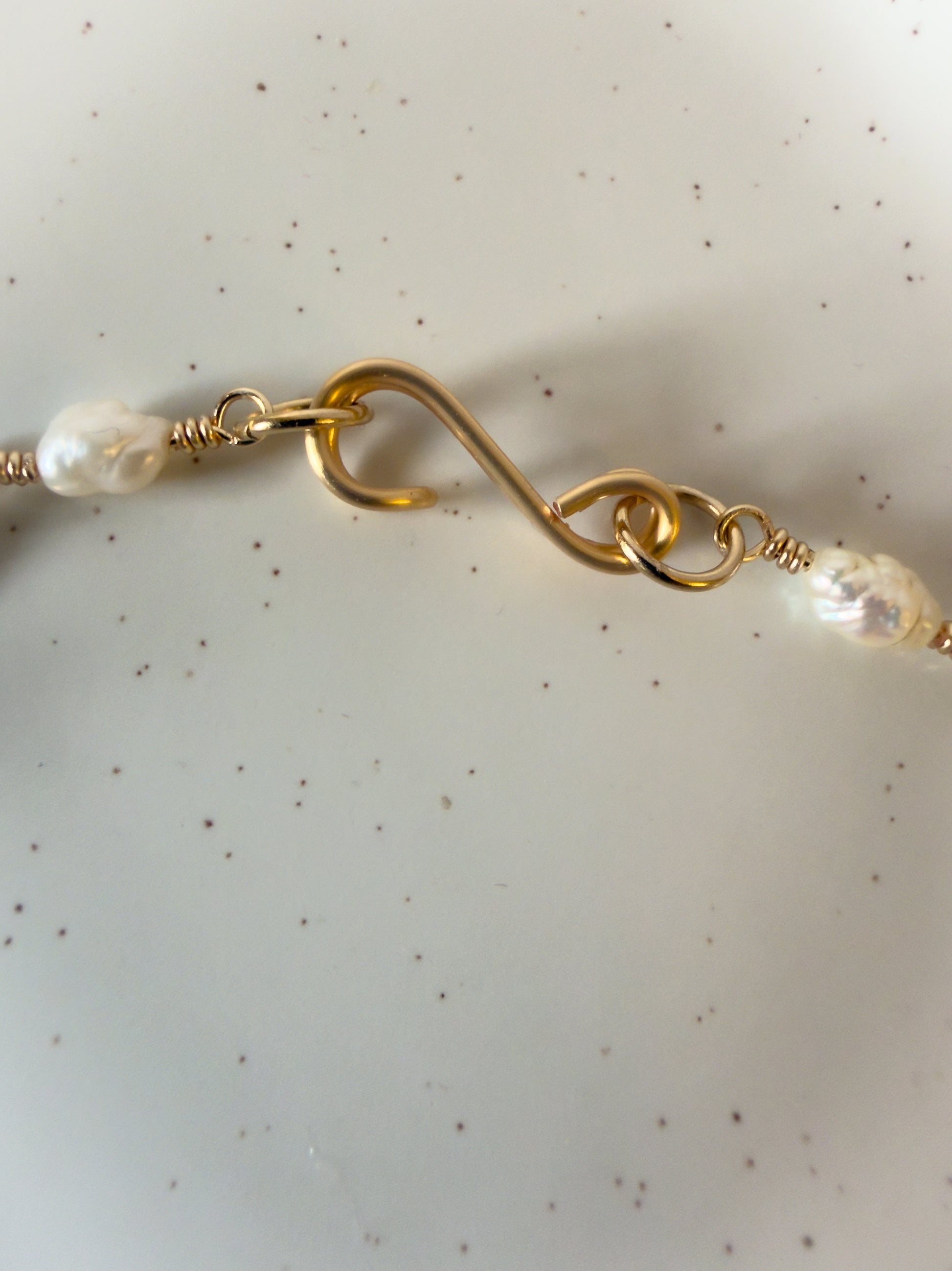 Close-up of a handmade gold necklace closure with pearl beads on a speckled beige background