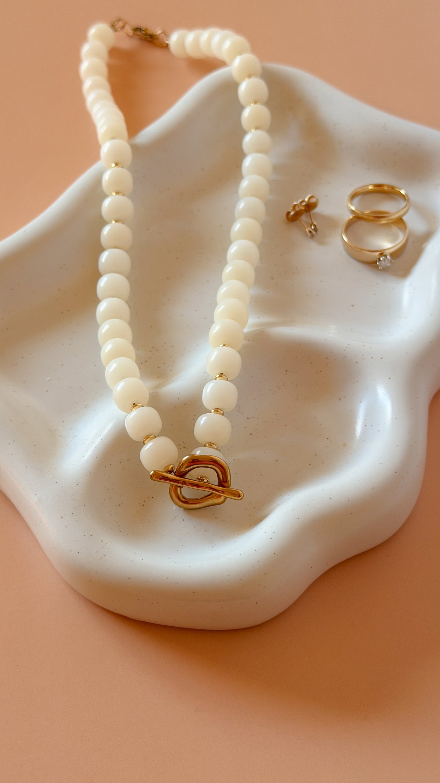 A cream-colored beaded necklace with a gold heart-shaped toggle clasp, displayed on a white ceramic dish alongside two gold rings against a peach background.