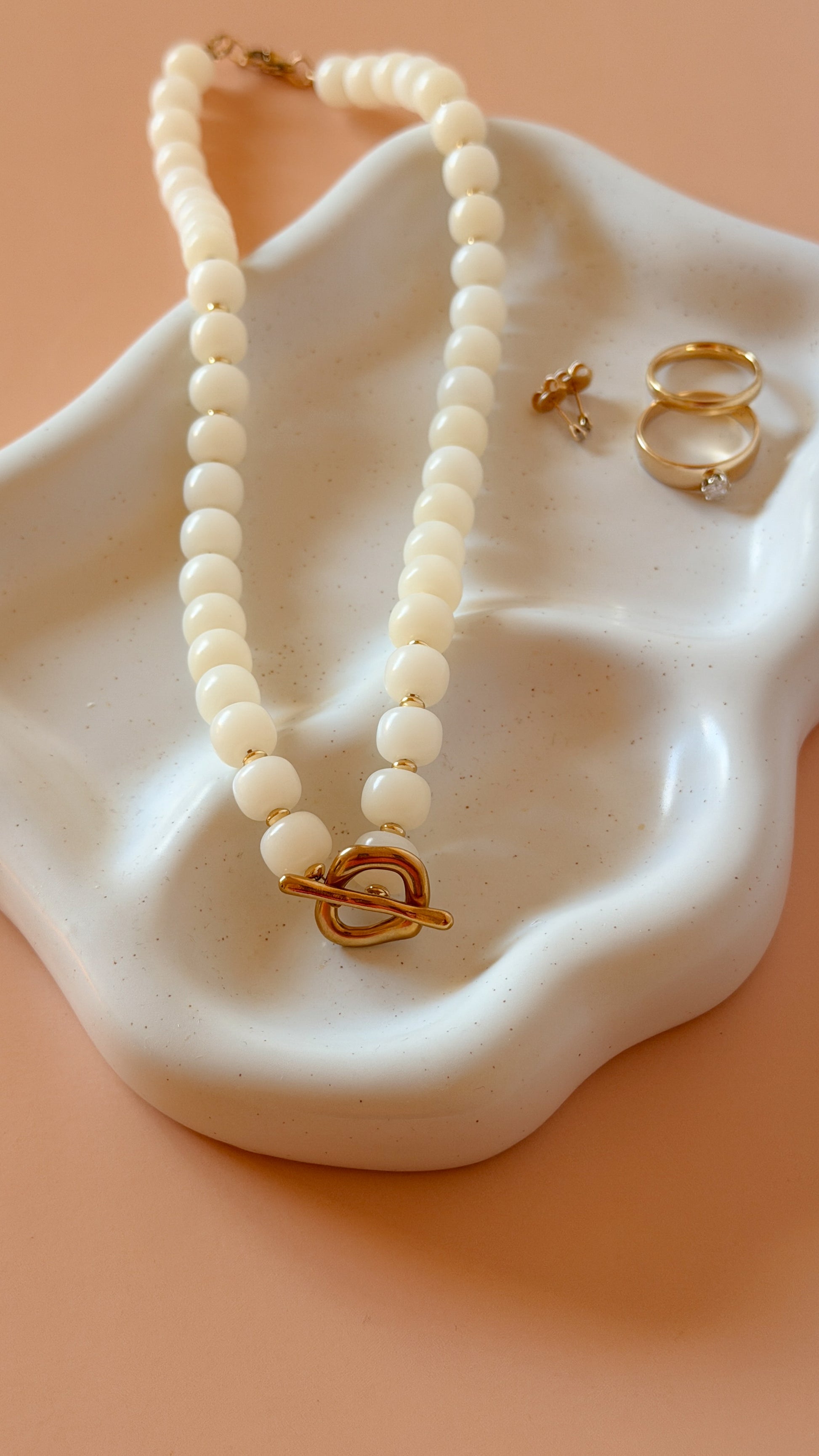 A cream-colored beaded necklace with a gold heart-shaped toggle clasp, displayed on a white ceramic dish alongside two gold rings against a peach background.