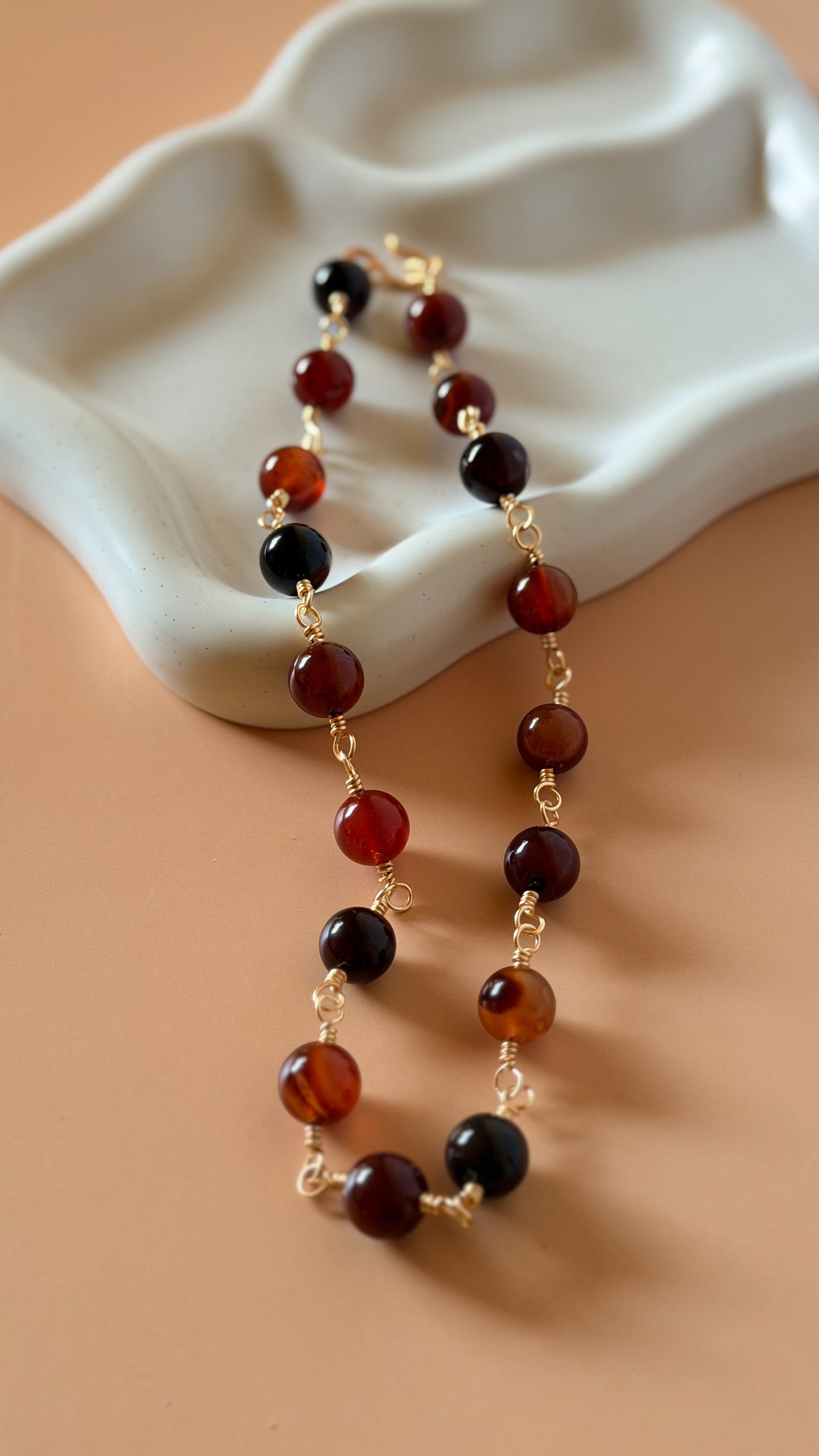 A beaded necklace with alternating translucent amber-brown and glossy black round beads, separated by small gold chain links, draped over a white ceramic dish on a warm beige background.