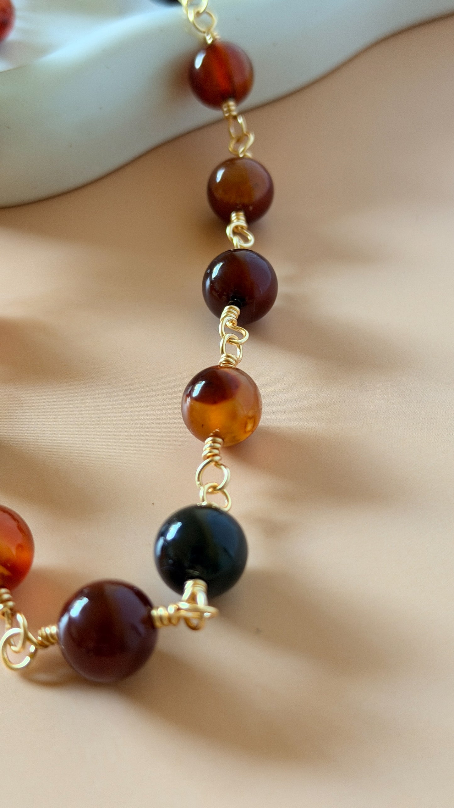 A close-up of a beaded necklace featuring polished amber-brown and dark black round beads separated by small gold chain links, draped over a neutral beige surface.