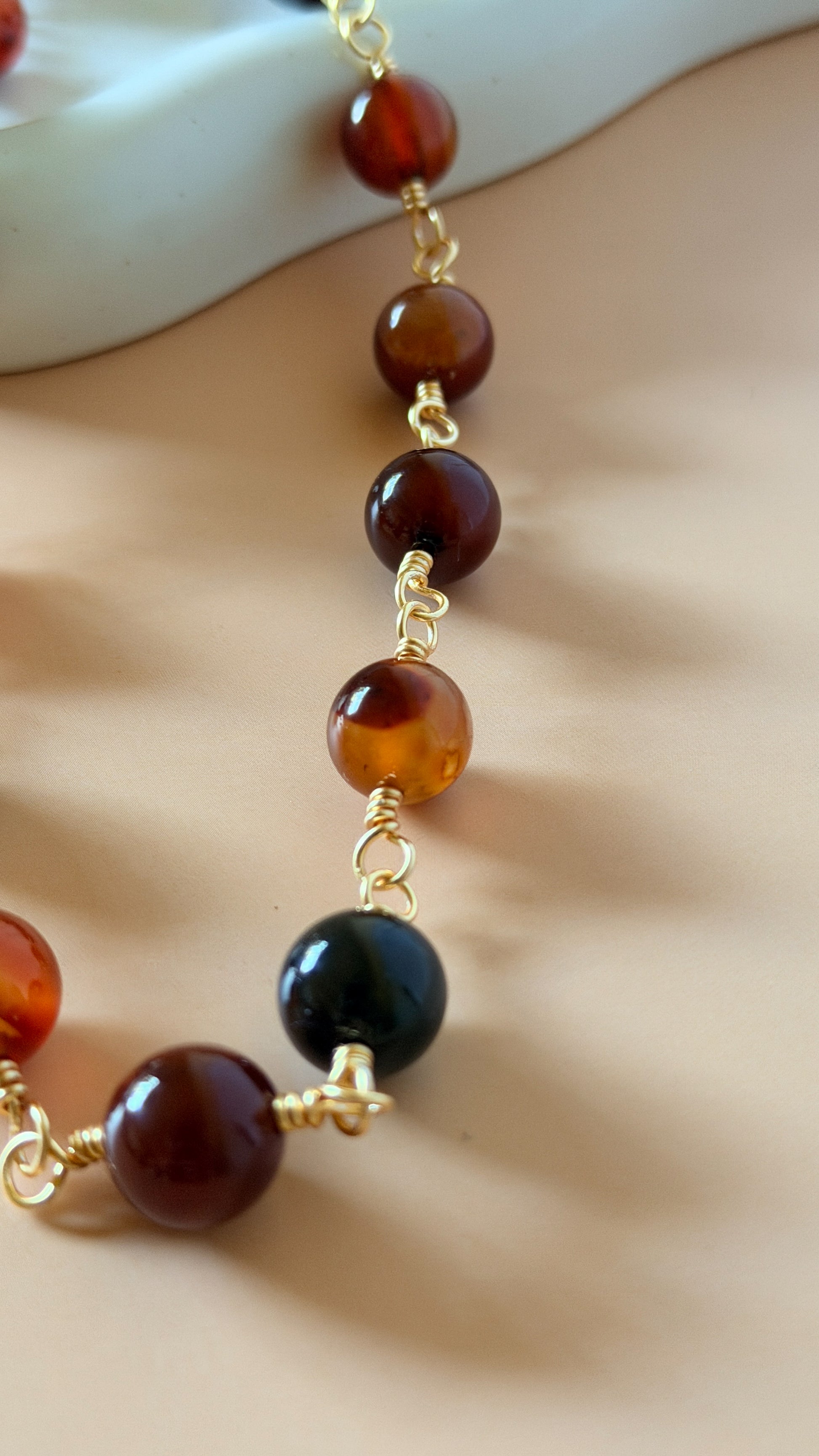 A close-up of a beaded necklace featuring polished amber-brown and dark black round beads separated by small gold chain links, draped over a neutral beige surface.