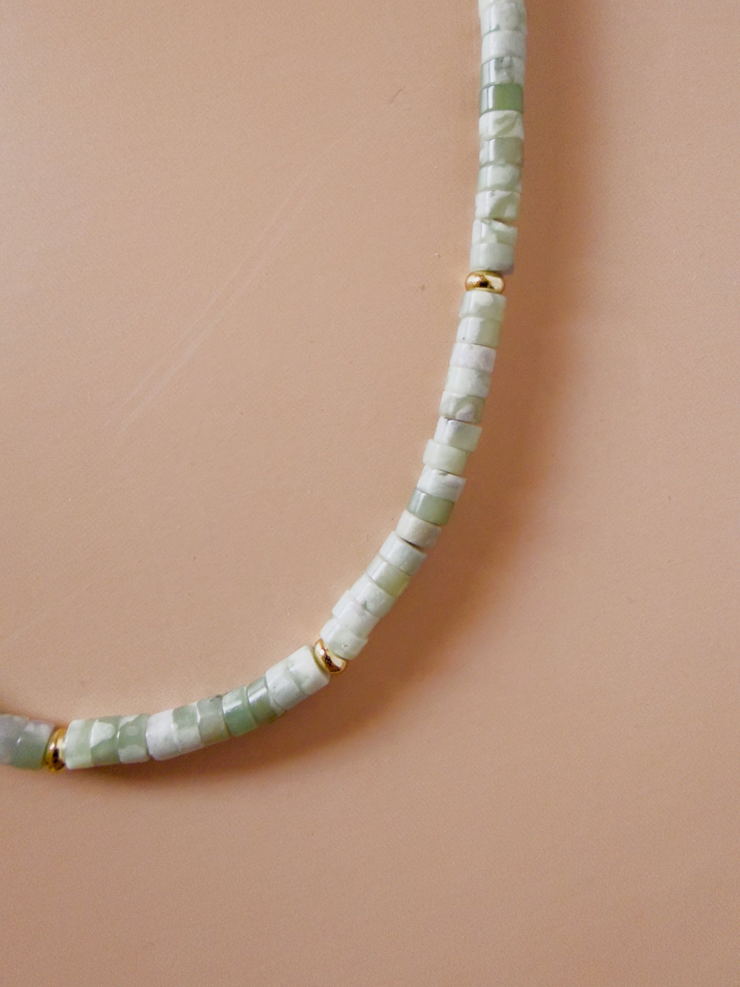 A close-up of a beaded necklace made of light green and white faceted beads separated by small gold spacer rings, laid on a warm peach background.