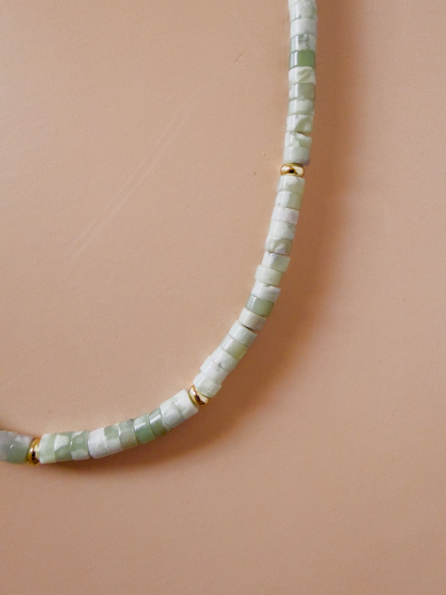 A close-up of a beaded necklace made of light green and white faceted beads separated by small gold spacer rings, laid on a warm peach background.