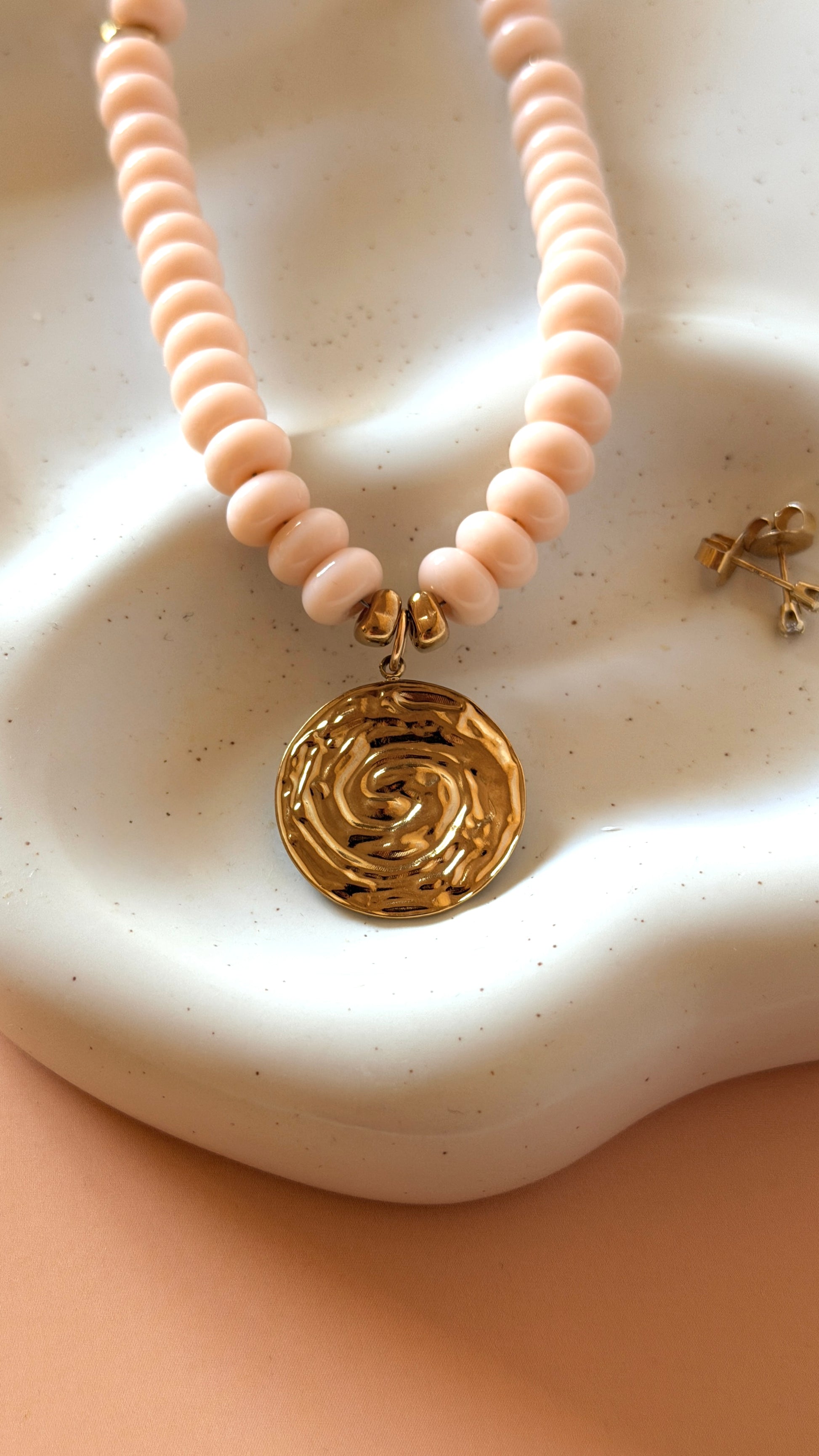 A close-up of a necklace with pale blush pink round beads and a textured gold pendant, displayed on a white ceramic dish against a peach background.