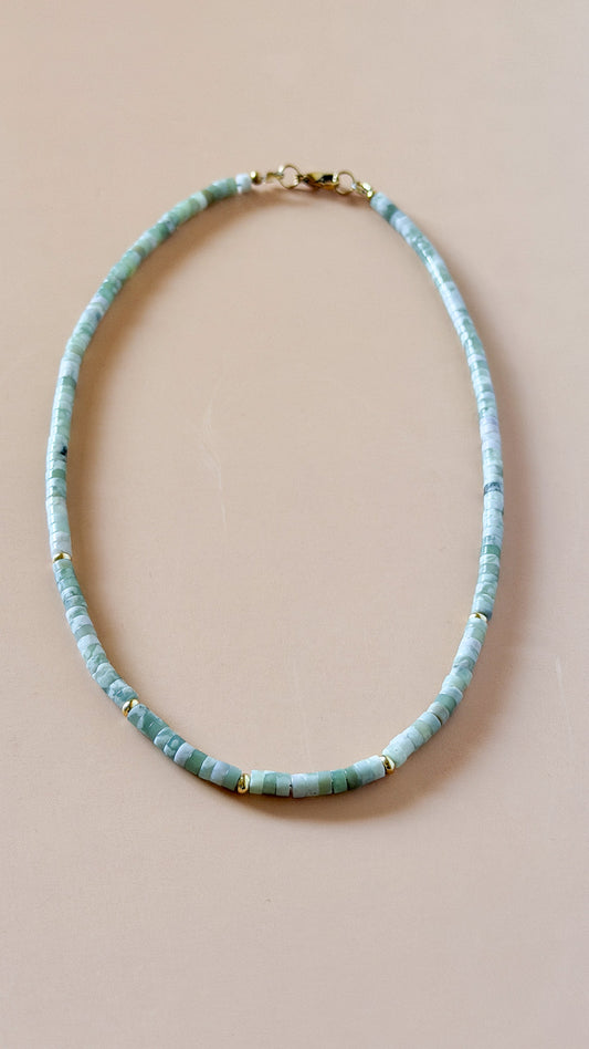 A beaded necklace made of small light blue-green stone beads with tiny gold spacer accents and a gold clasp, displayed on a solid beige background.