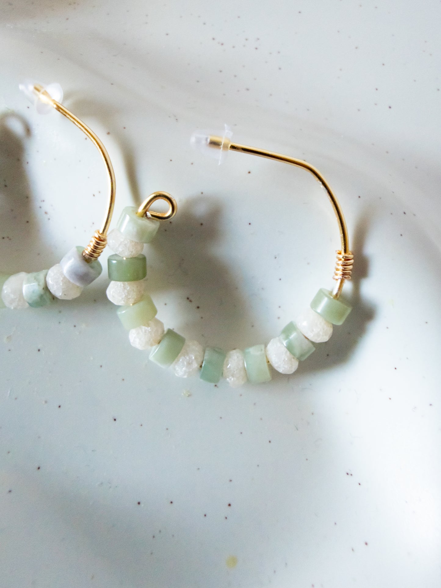 Gold hoop earrings with dangling beaded strands featuring light green and white stones, displayed on a light speckled surface.