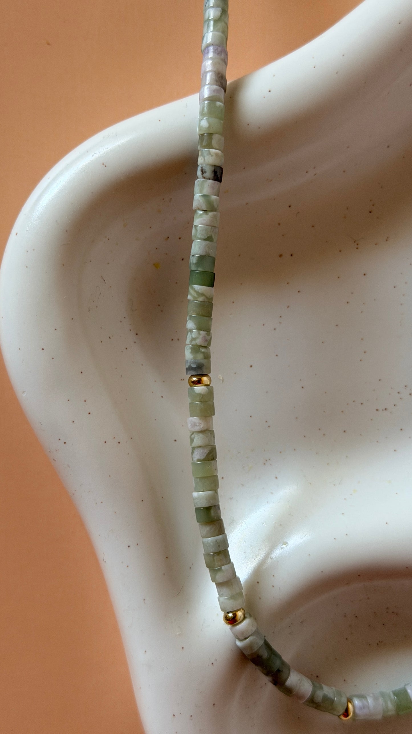 A close-up of a beaded necklace draped over a white stone jewelry display bust. The necklace consists of small, chunky stone beads in shades of sage green, light gray, and white, accented with a few small gold spacer beads.