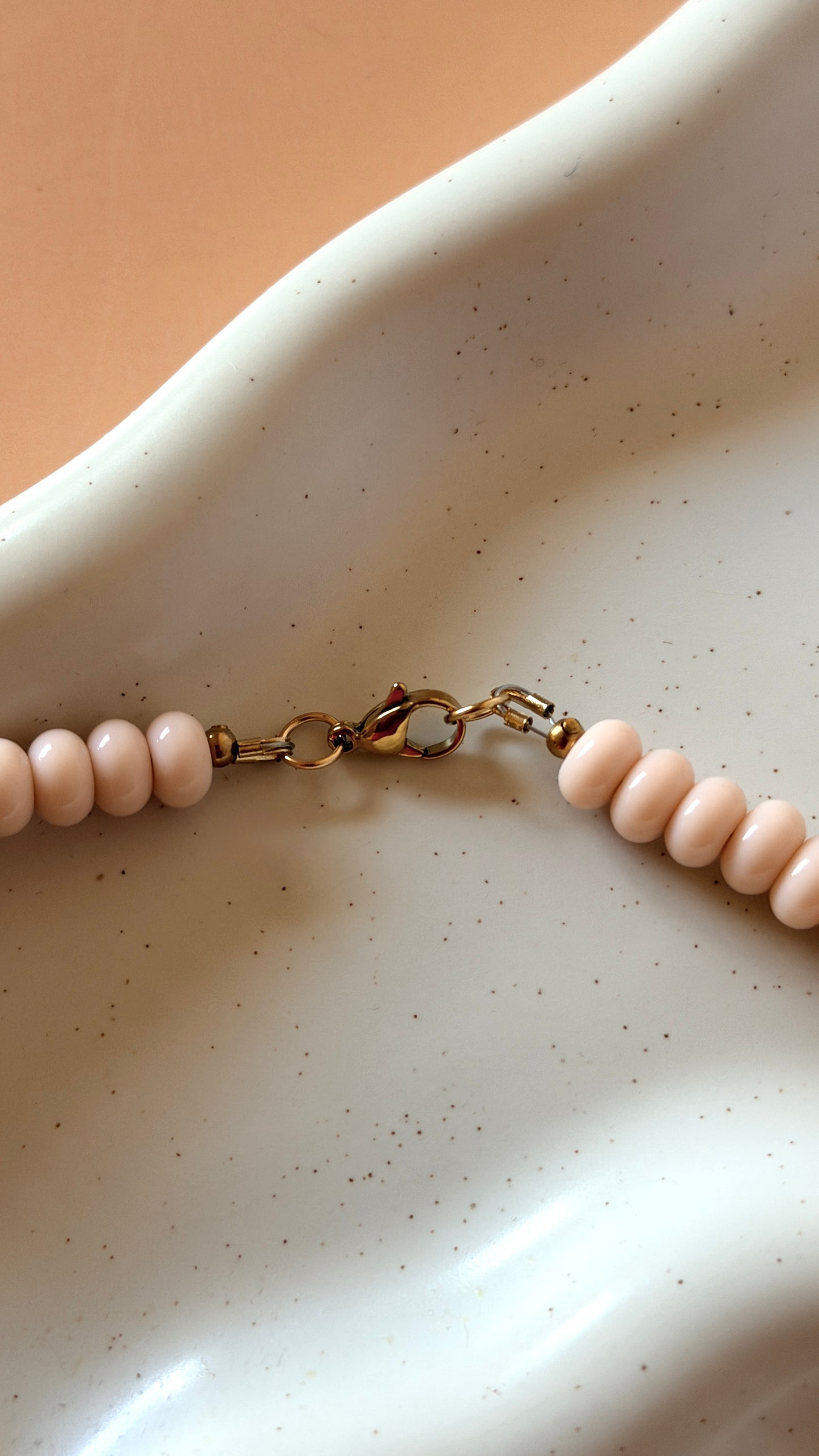 A close-up of a beaded necklace with pale pink round beads and a gold clasp, resting on a white speckled ceramic dish against a warm beige background.