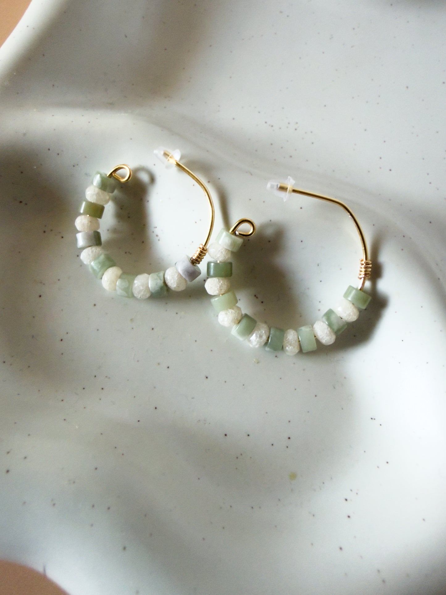 Gold hoop earrings with small green and white beads, resting on a white speckled ceramic dish.