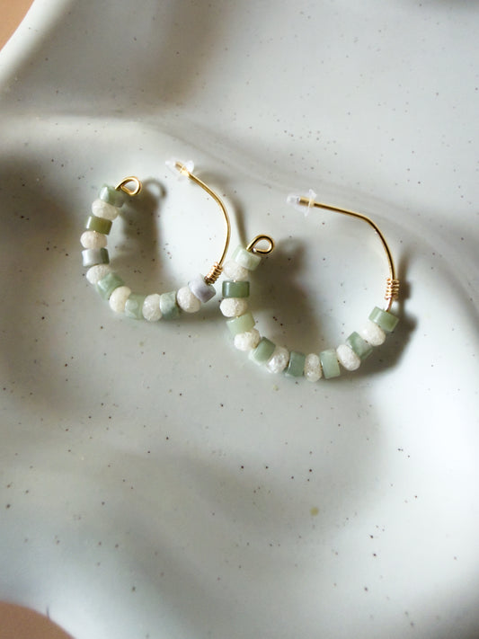Gold hoop earrings with small green and white beads, resting on a white speckled ceramic dish.