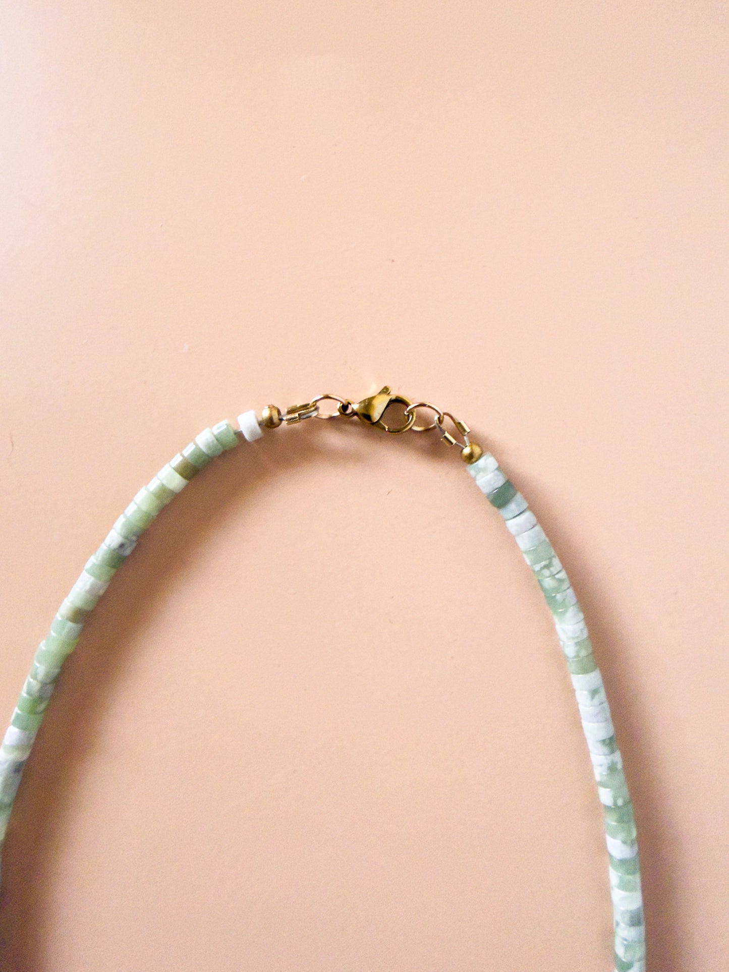 A beaded necklace made of light green and white marbled beads with a gold clasp, displayed on a solid peach-pink background.