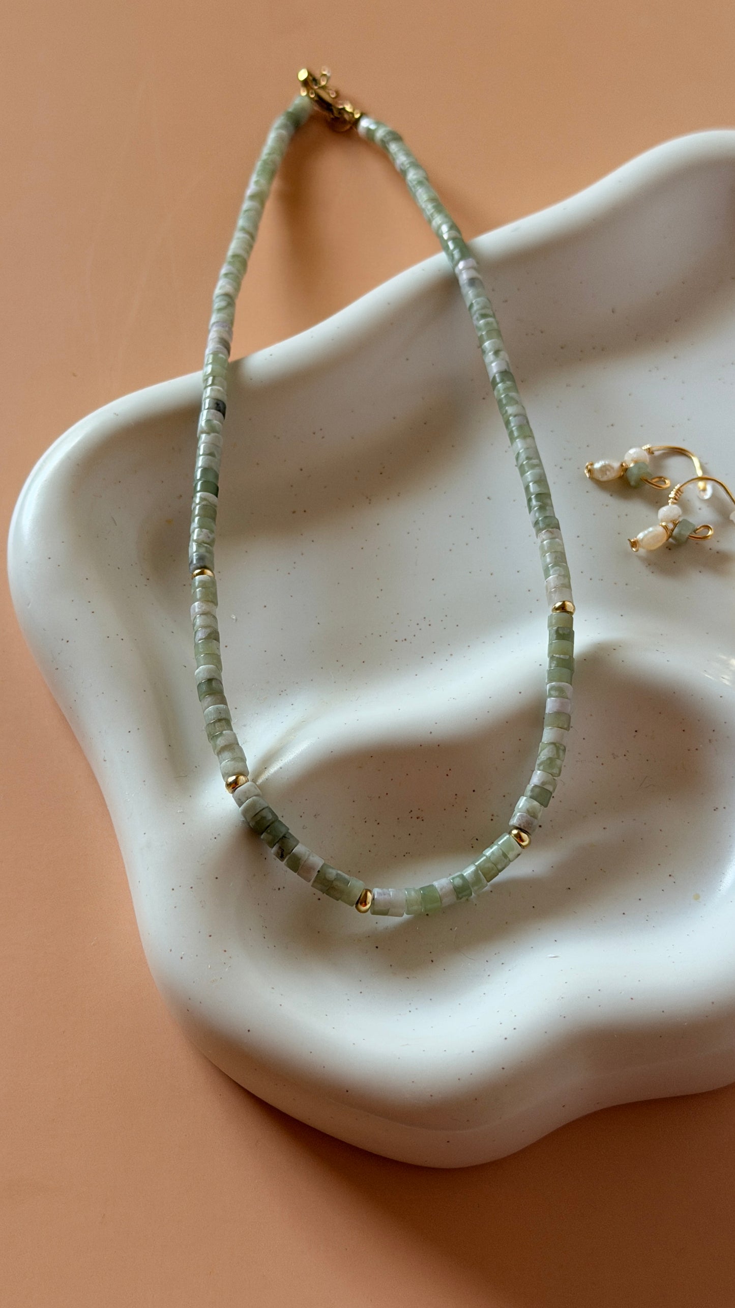 A beaded necklace made of light green stone beads with small gold spacer beads and a gold clasp, displayed on a white ceramic jewelry stand against a peach background.