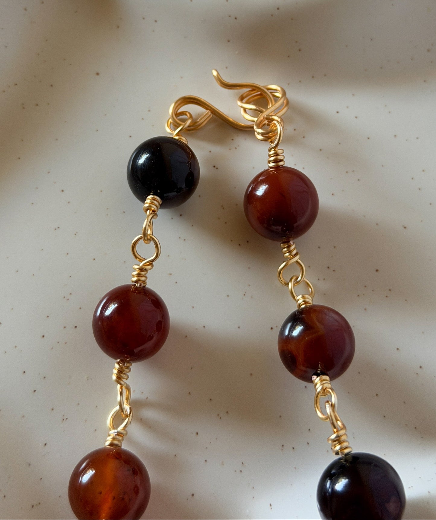 A pair of gold-tone drop earrings with glossy dark amber and black beads, resting on a textured white ceramic surface.