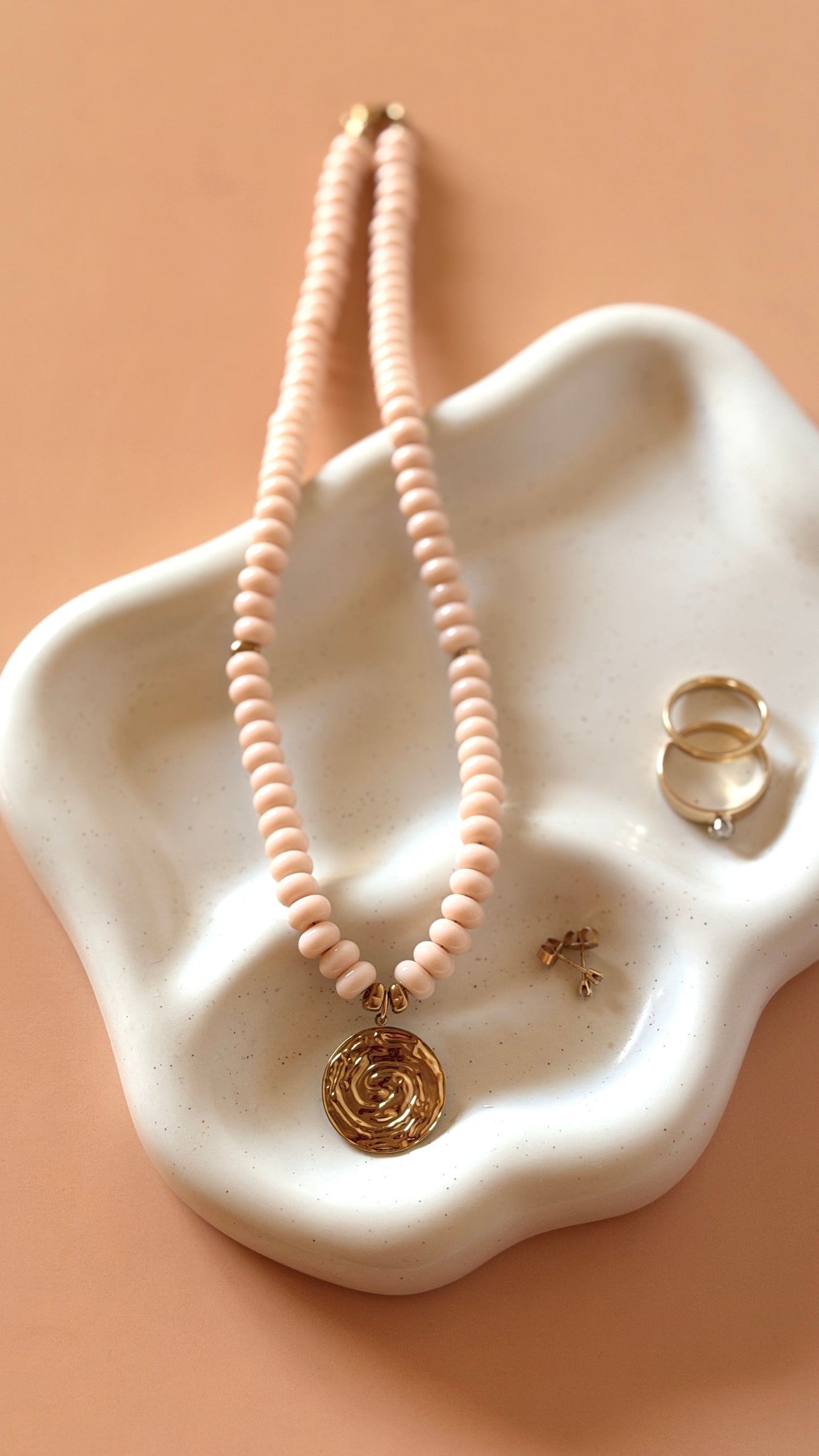 A pale pink beaded necklace with a gold rose pendant, displayed on a white ceramic dish alongside two gold rings against a peach background.