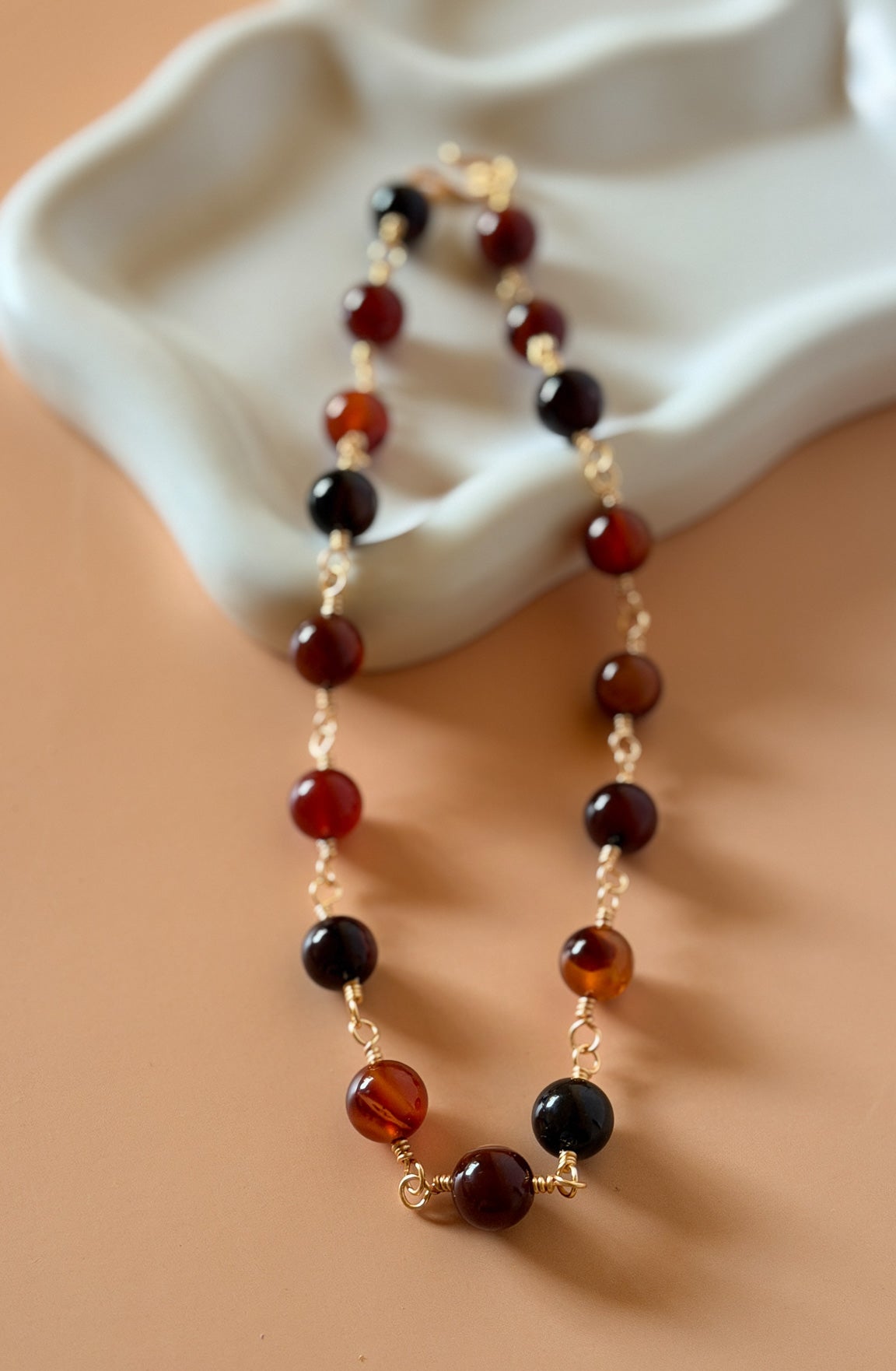 A beaded necklace with alternating translucent amber-brown and glossy black round beads, separated by small gold chain links, displayed on a warm beige surface with a white sculptural prop.